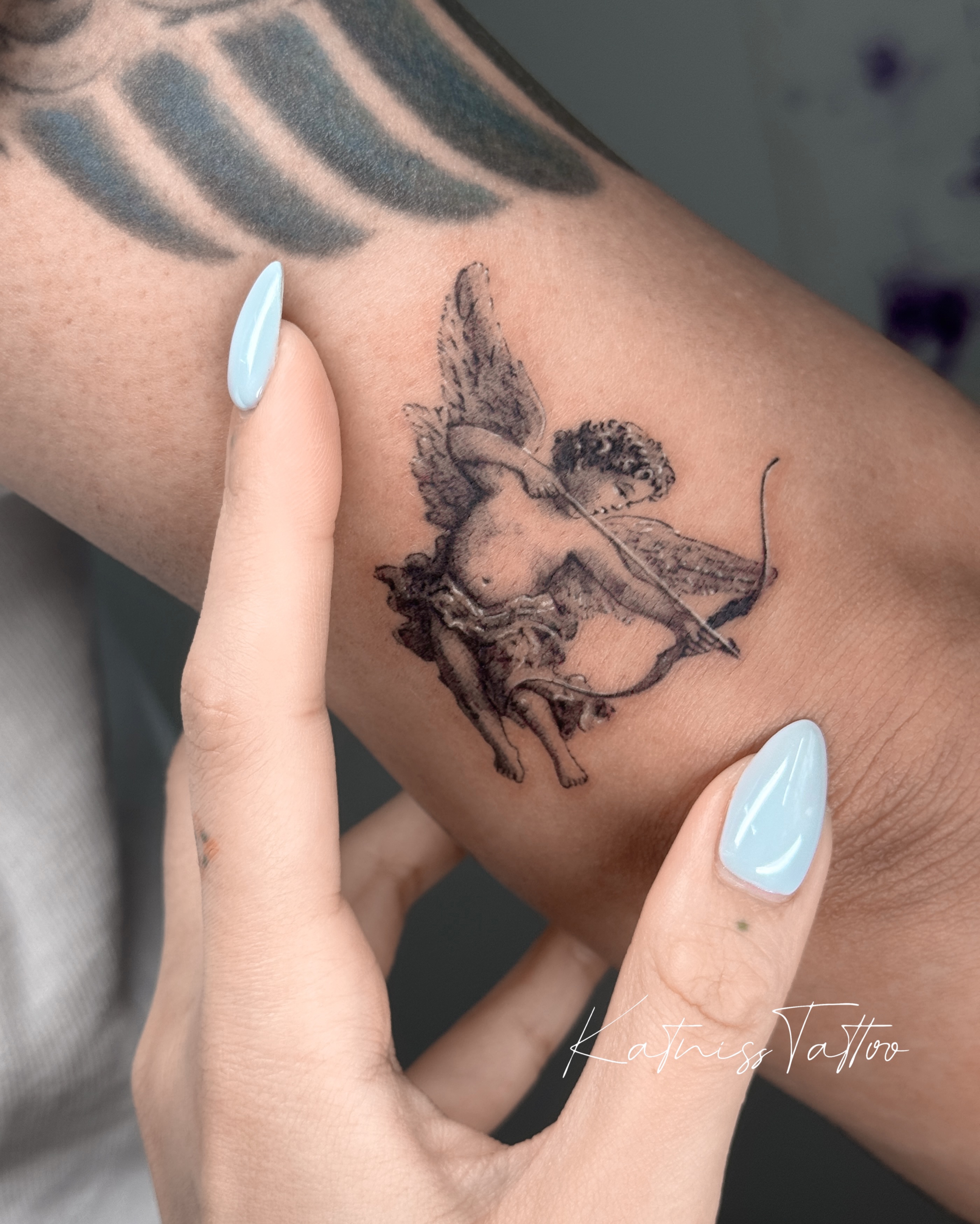 Cherub with bow black and grey tattoo on the forearm by 𝙆𝙖𝙩𝙣𝙞𝙨𝙨
