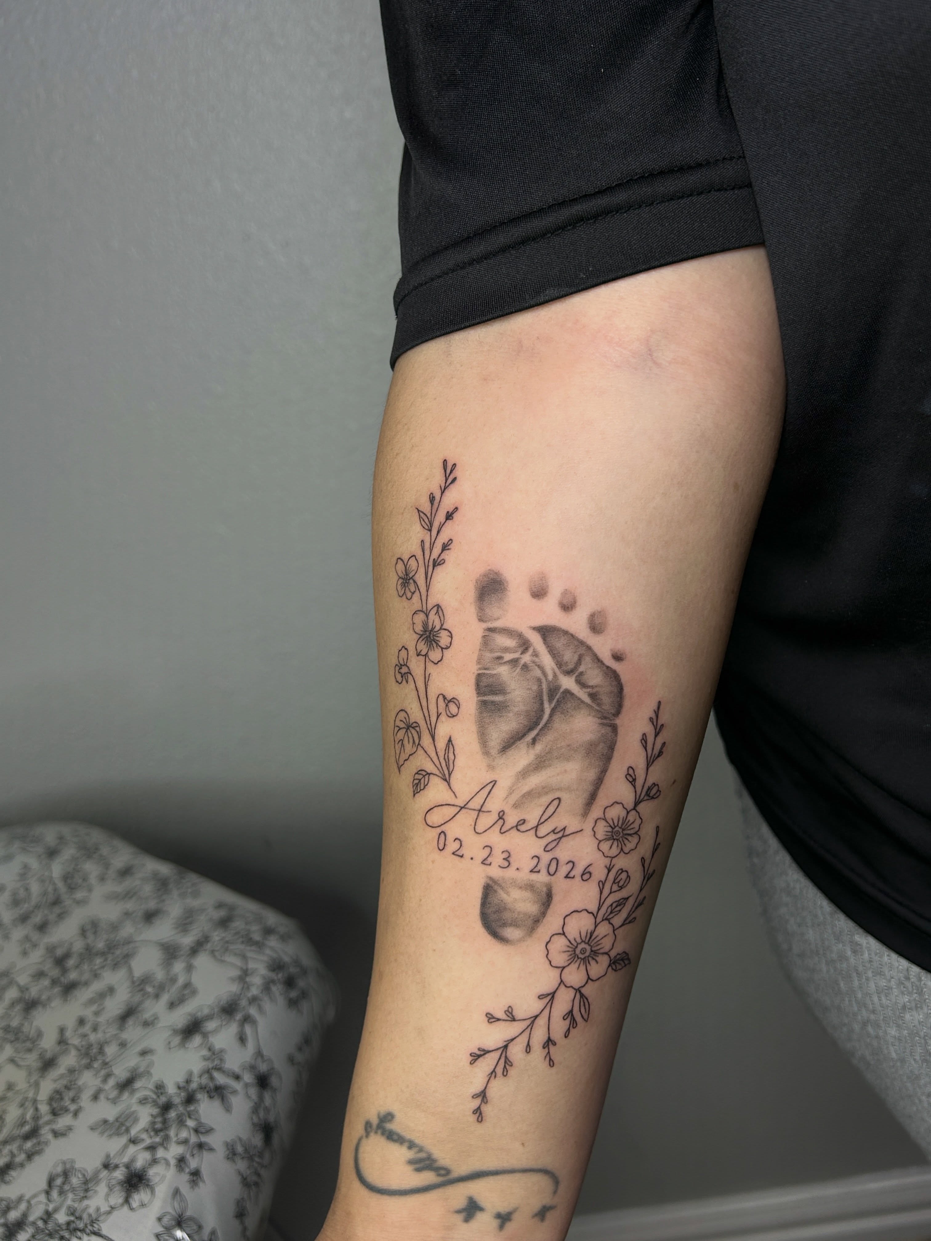 Baby footprint with floral elements realism tattoo on the forearm by @tattoosbyskyeee