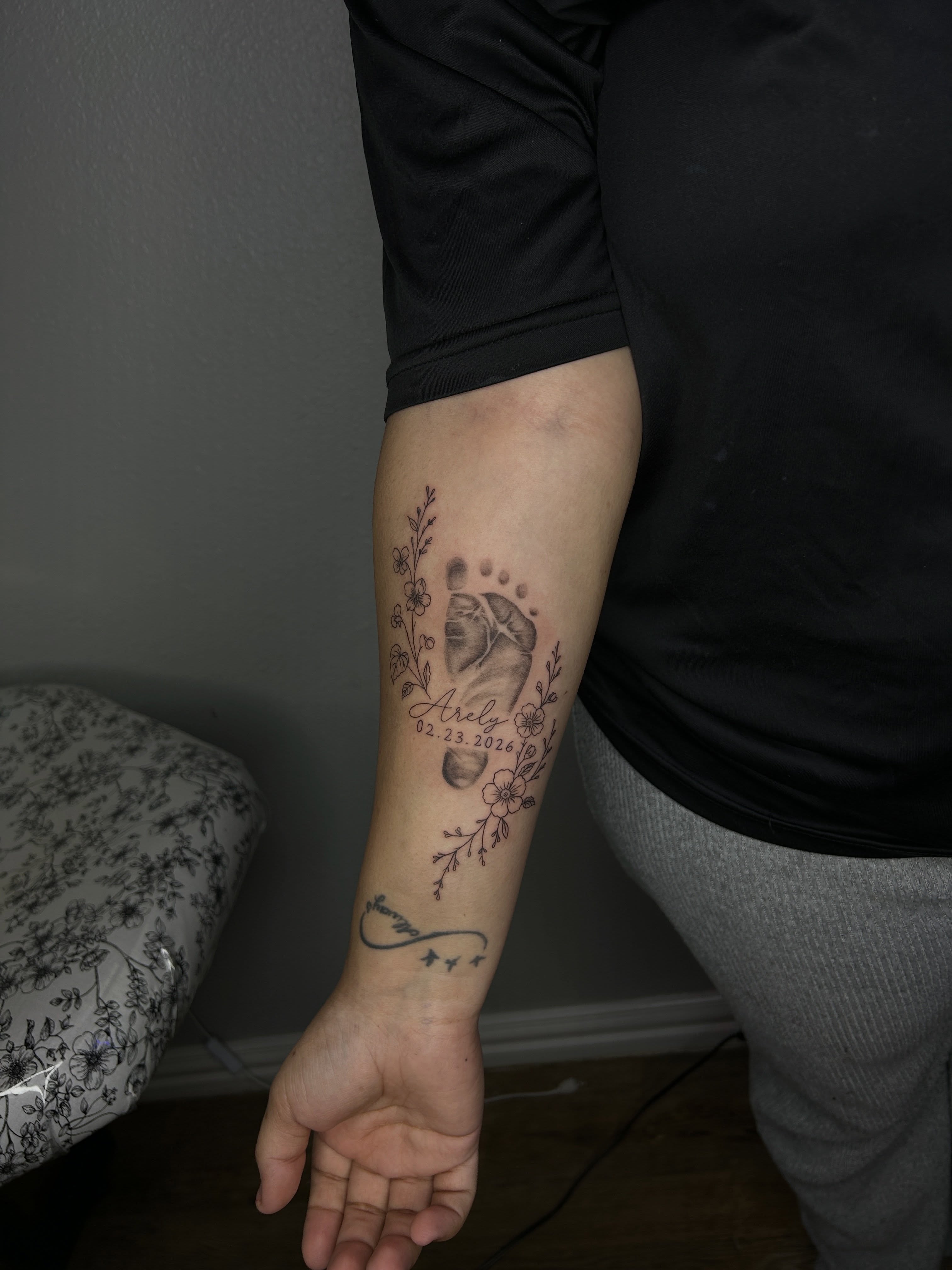 Baby footprints and floral design black and grey tattoo on the forearm by @tattoosbyskyeee