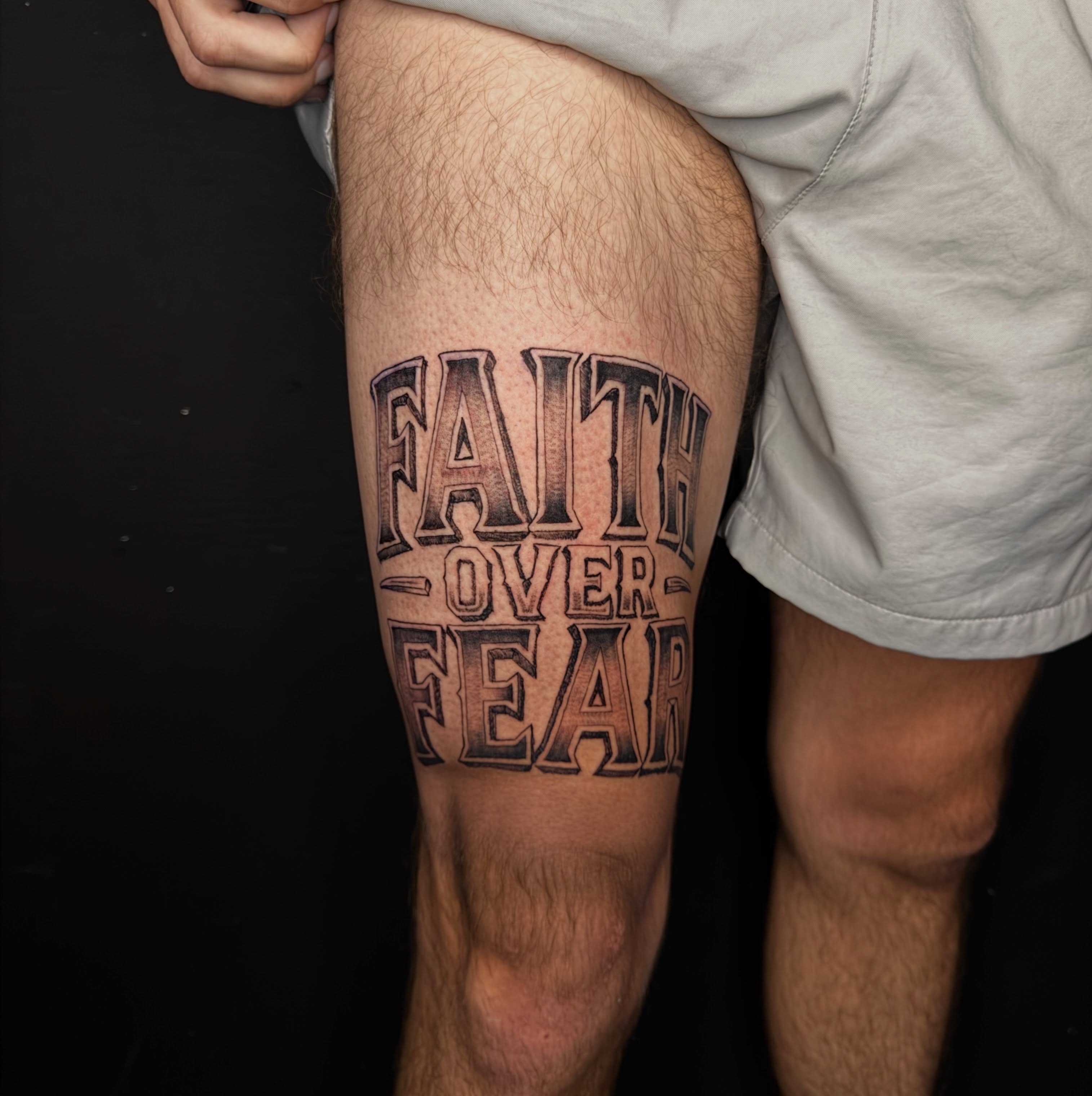 Faith over fear lettering script tattoo on the thigh by David Yancy III