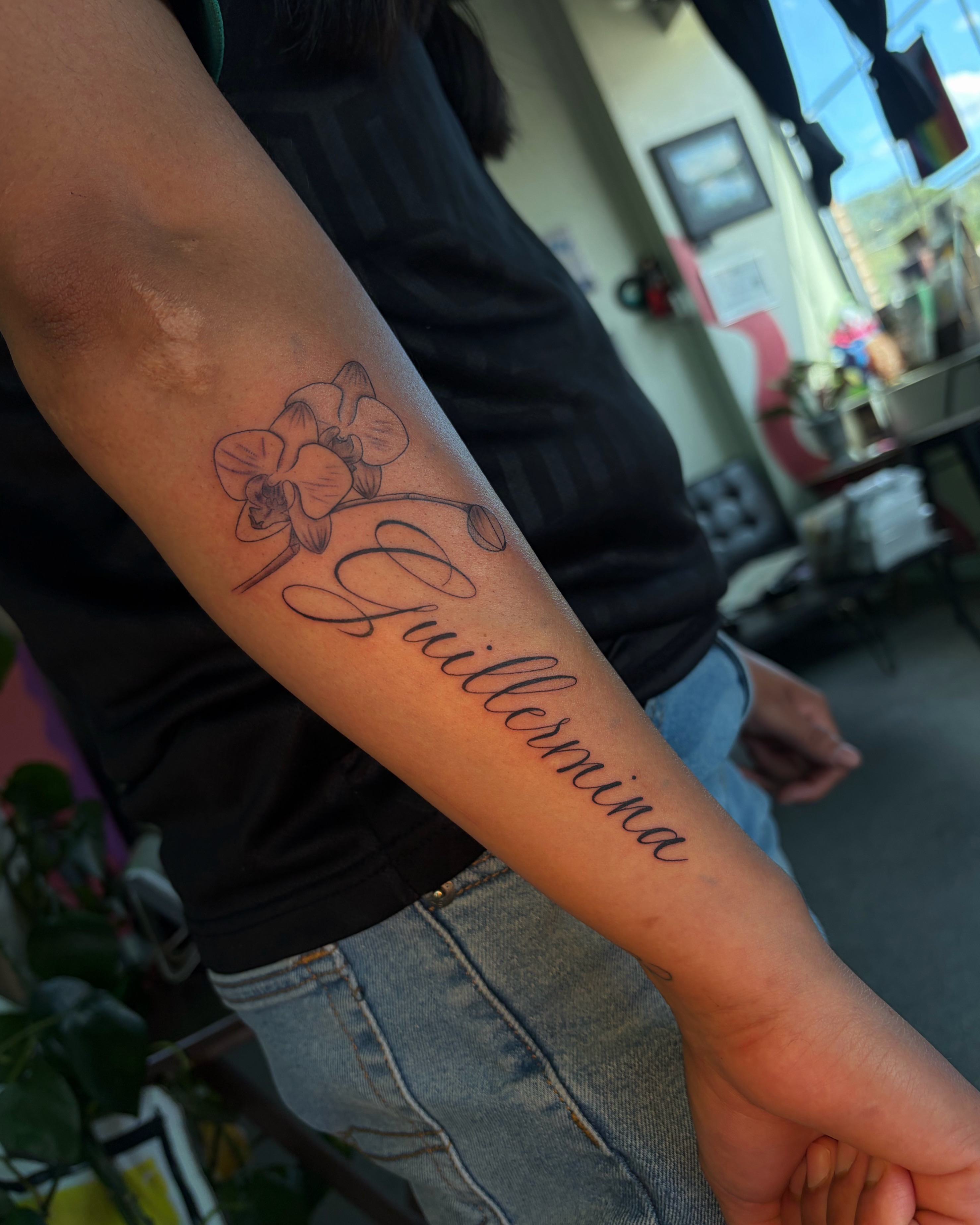 Orchid and name black and grey tattoo on the forearm by Daniela