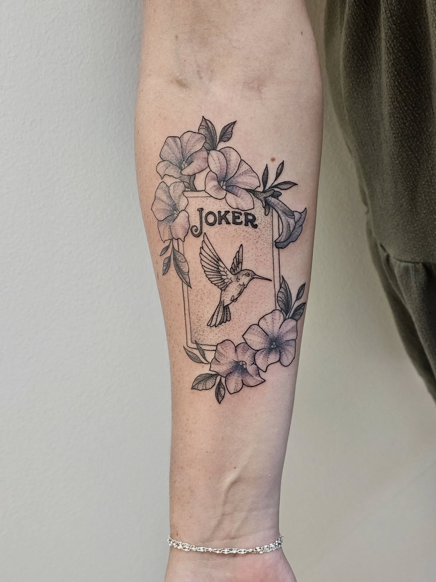 Joker card with hummingbird and flowers black and grey tattoo on the forearm by Kylie Lund