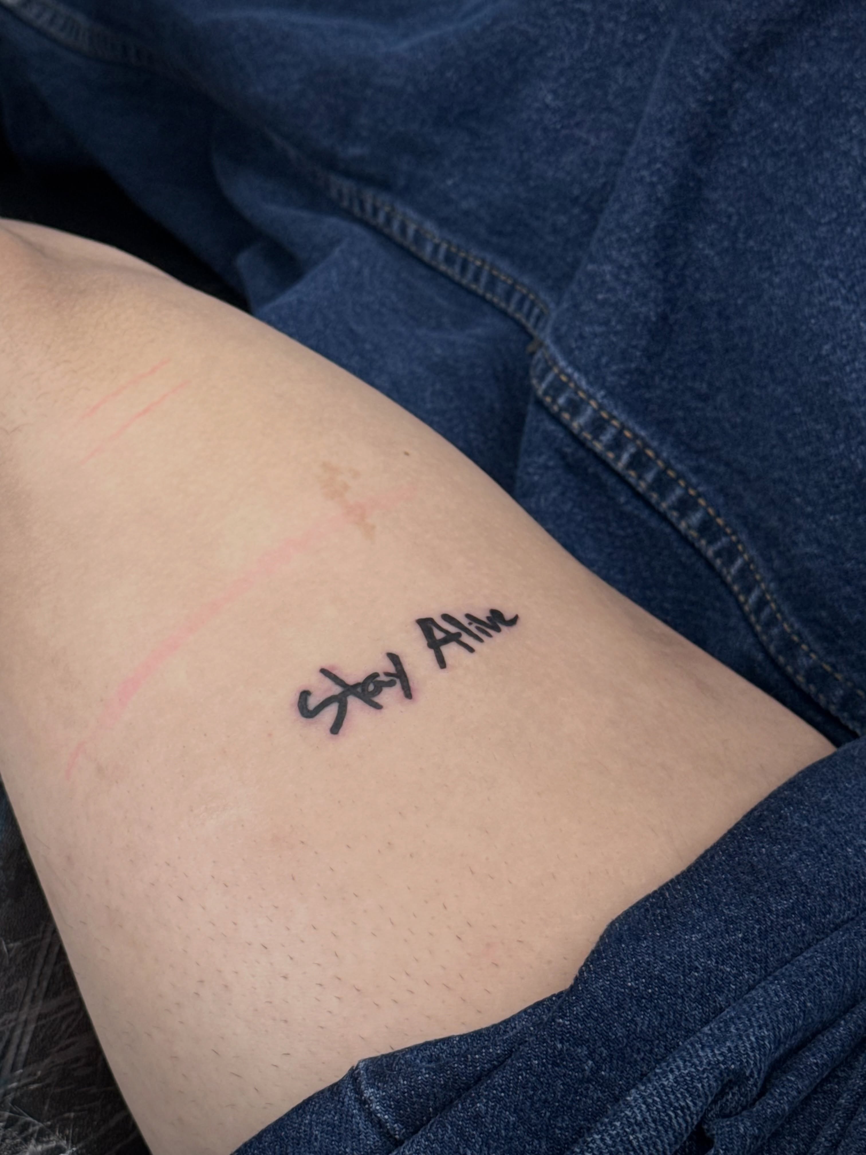 Stay Alive lettering script tattoo on the thigh by ⠀Greave