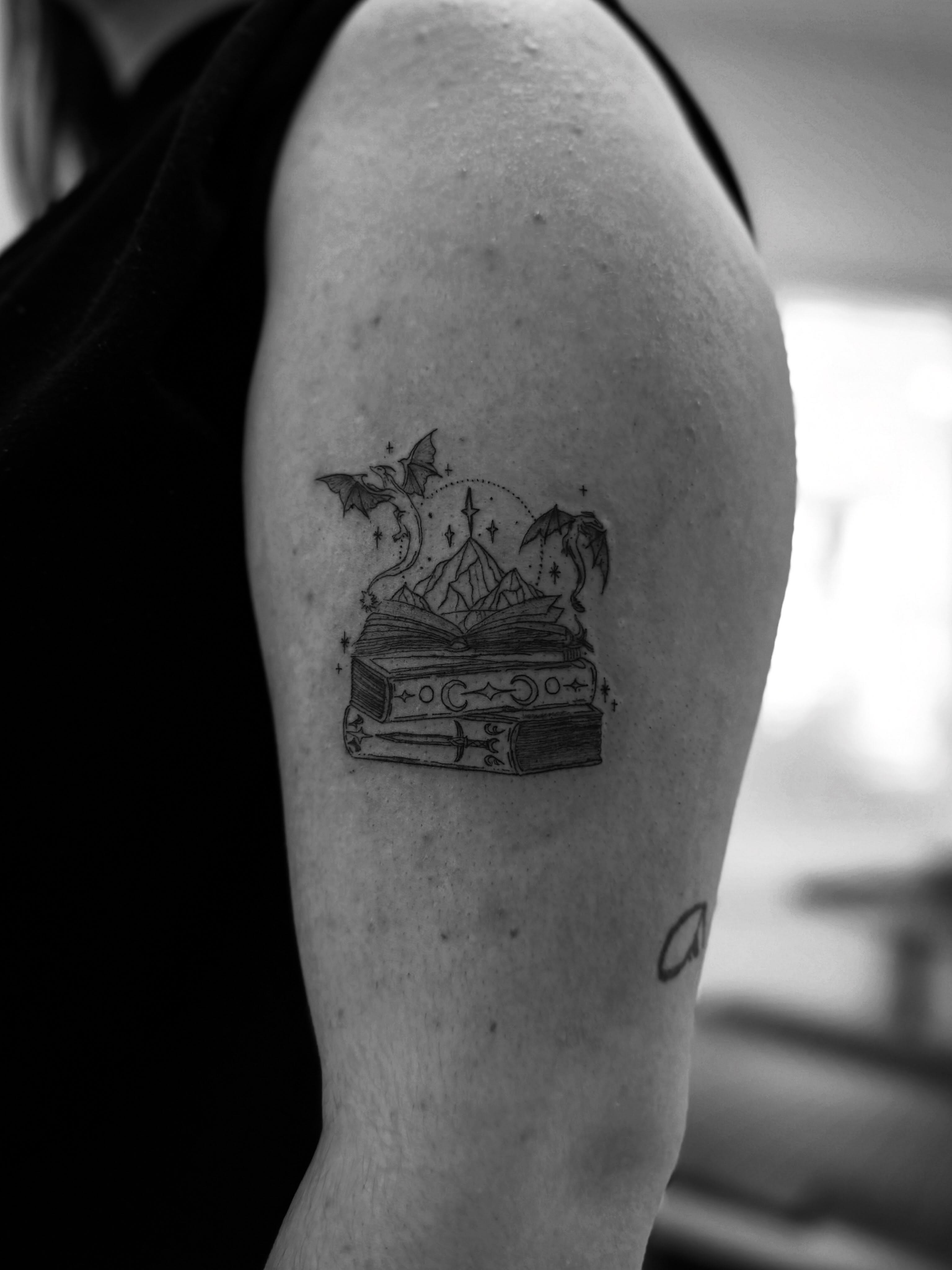 Book with magical elements black and grey tattoo on the upper arm by Taylor Johnson