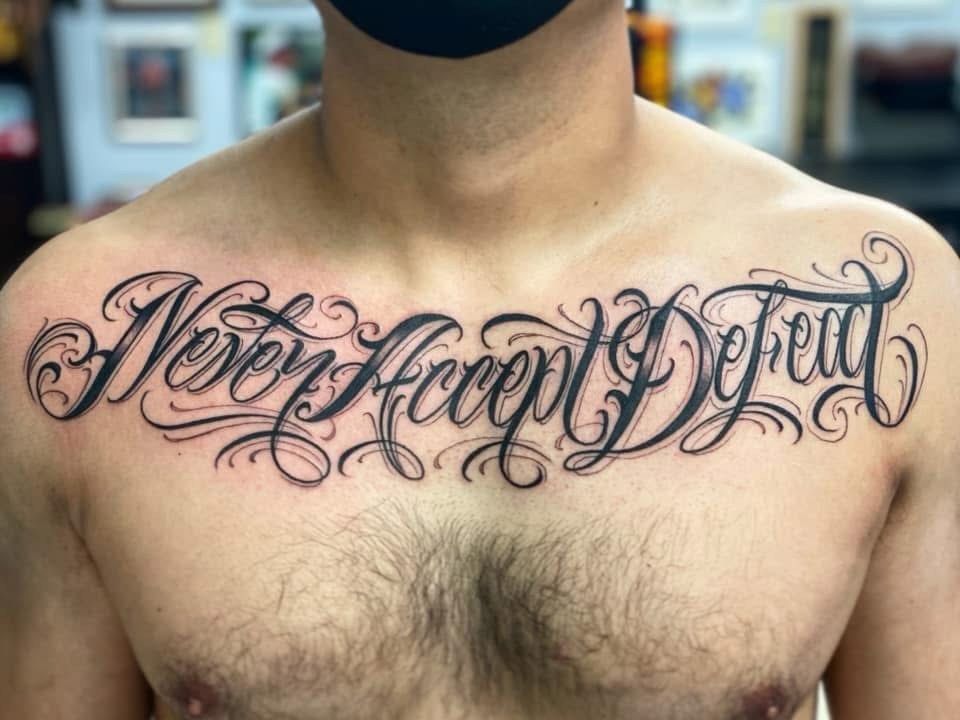 Never Accept Defeat lettering script tattoo on the chest by Lex Miranda