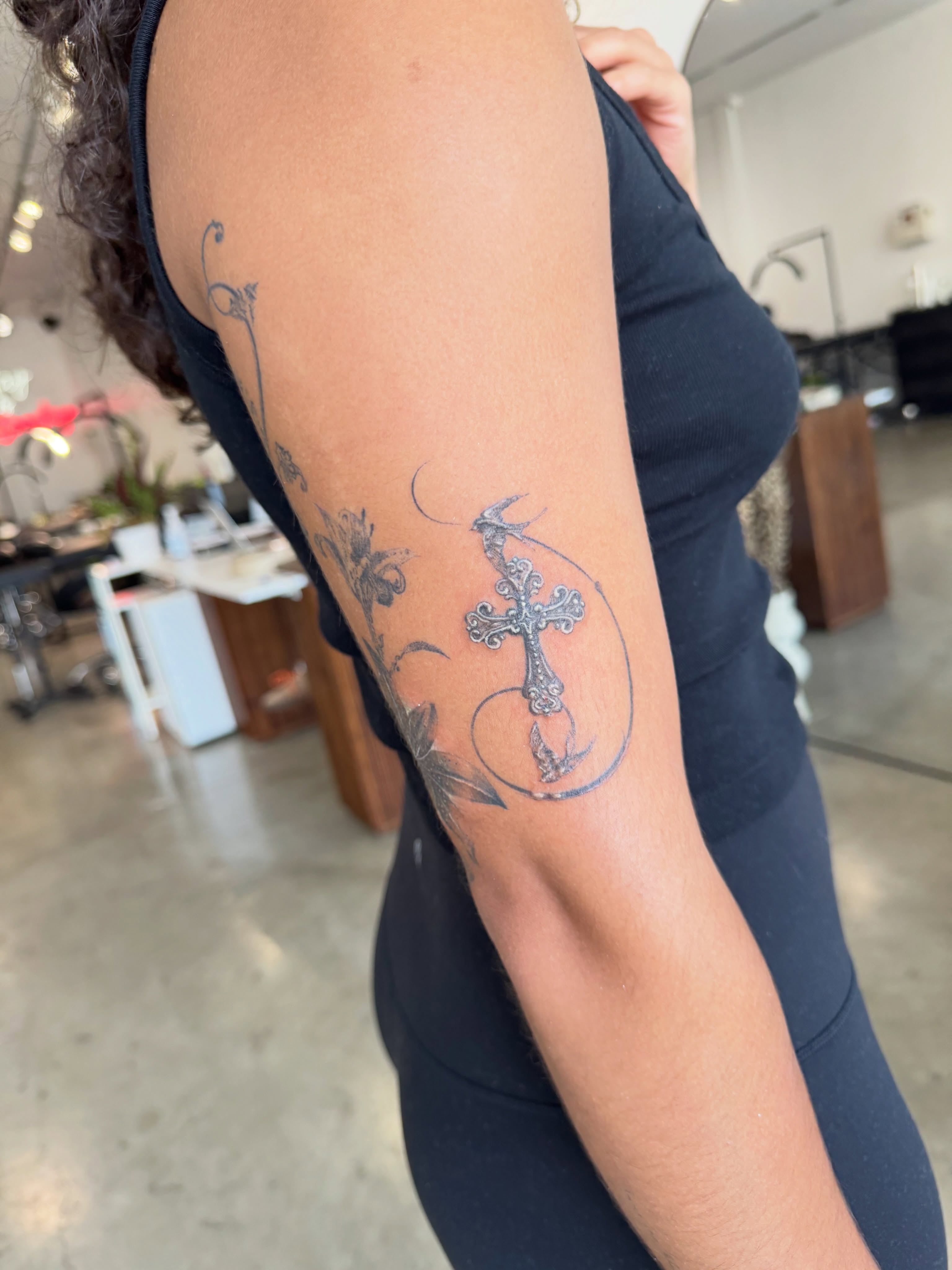Cross with floral elements black and grey tattoo on the forearm by Ava De La Torre