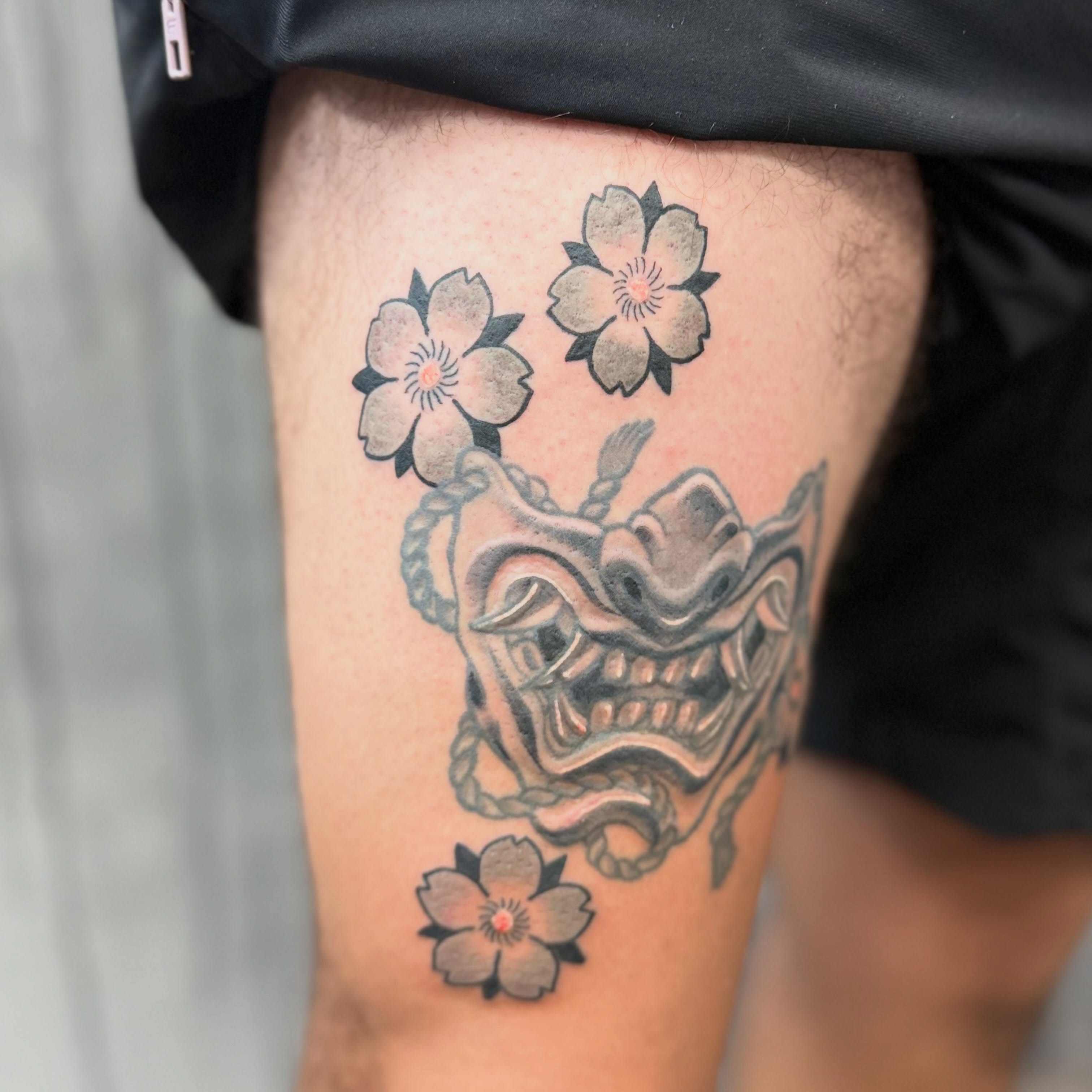 Mask with cherry blossoms black and grey tattoo on the thigh by Camilo