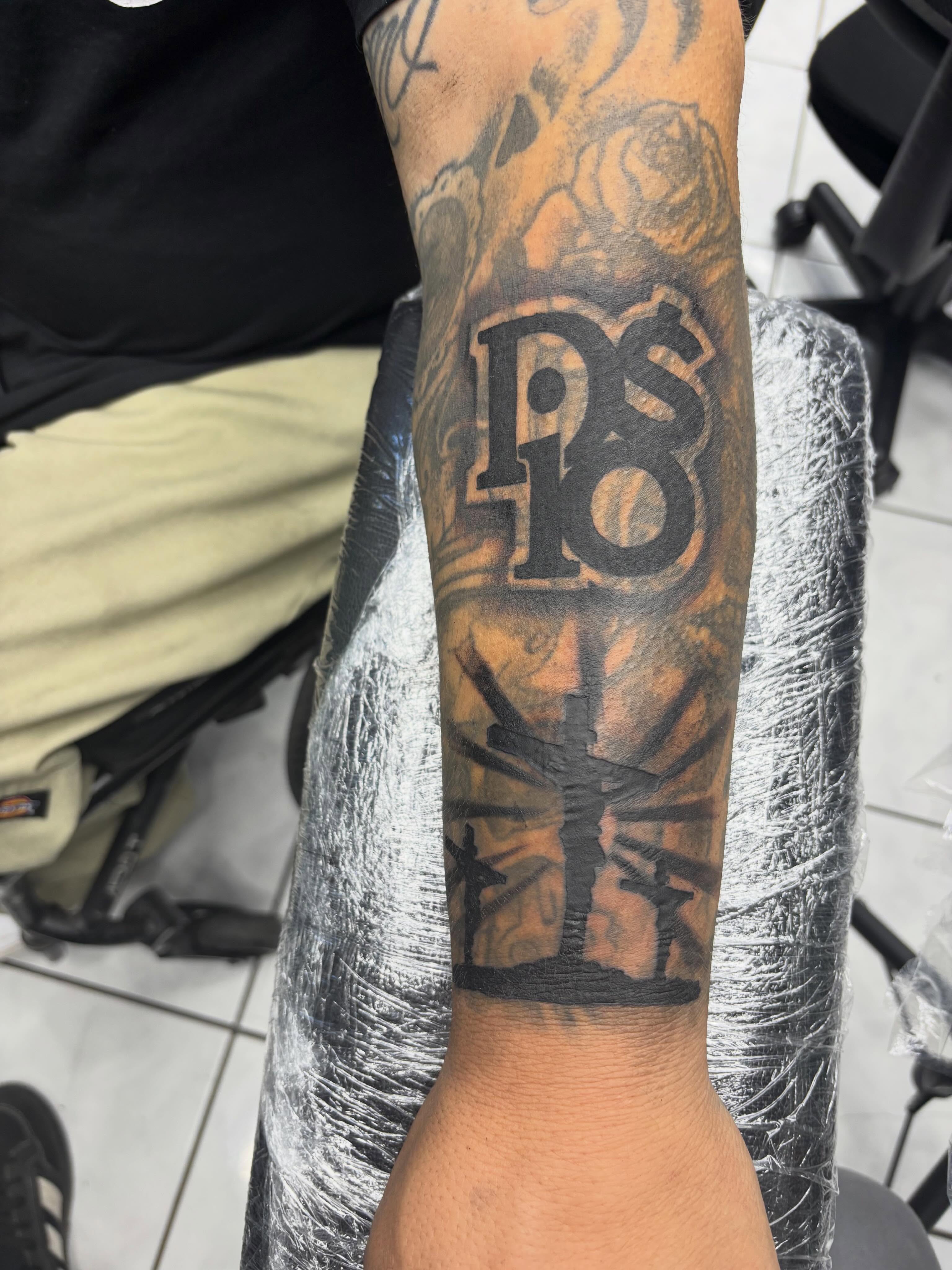 Cross with 'PS18' black and grey tattoo on the forearm by Shadows Tattoos