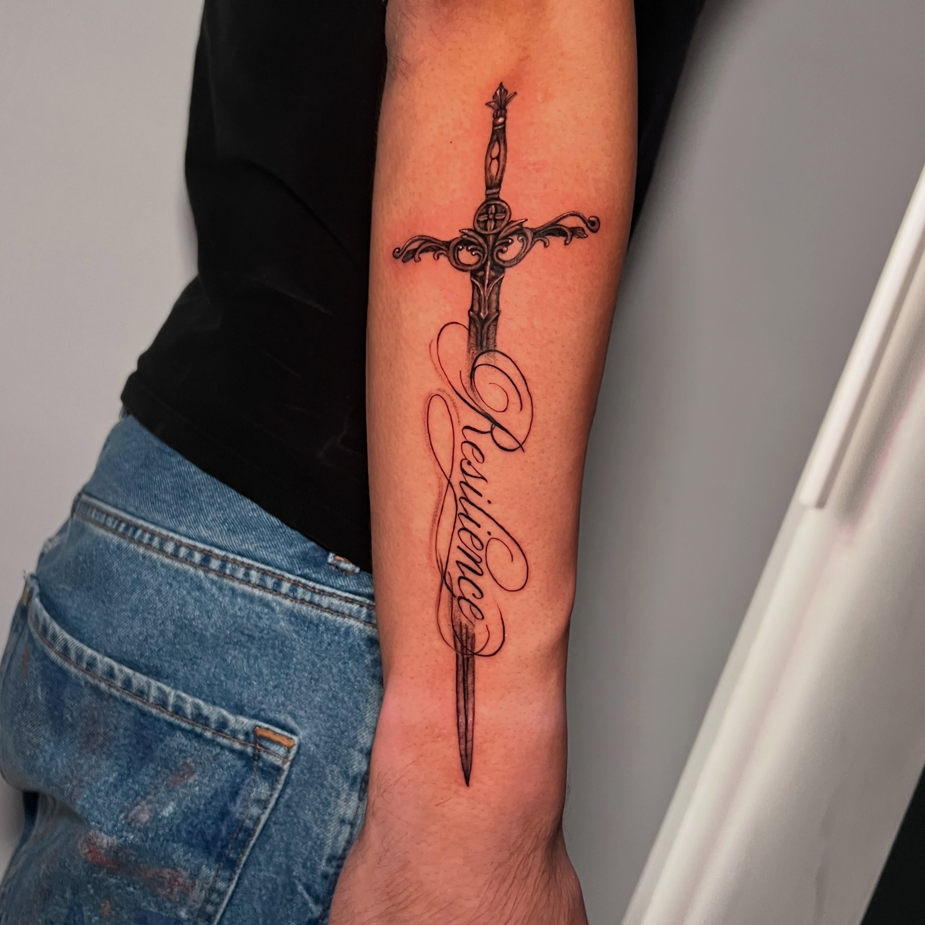 Sword and cursive text black and grey tattoo on the forearm by Eleana, North Miami, Florida