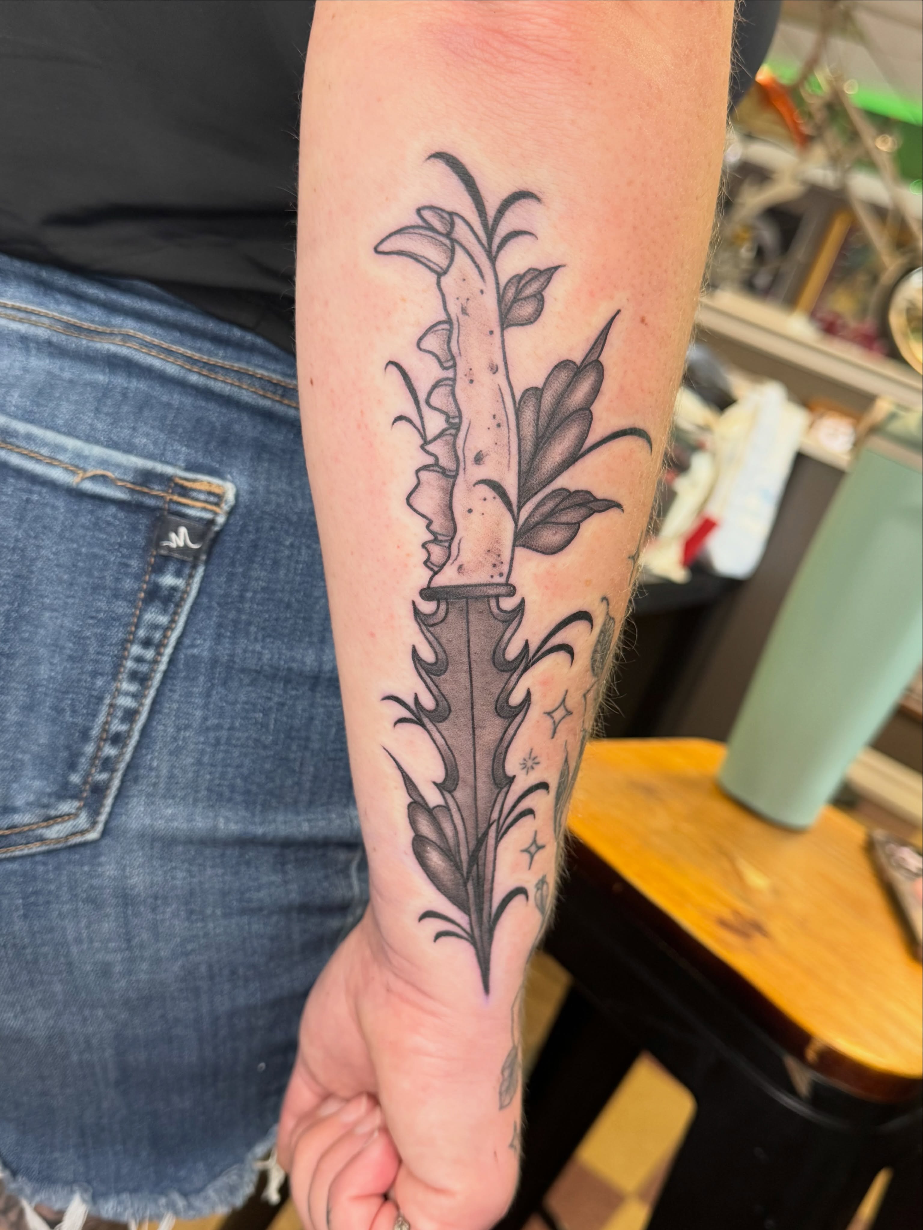 Knife and floral black and grey tattoo on the forearm by Cora Phelps