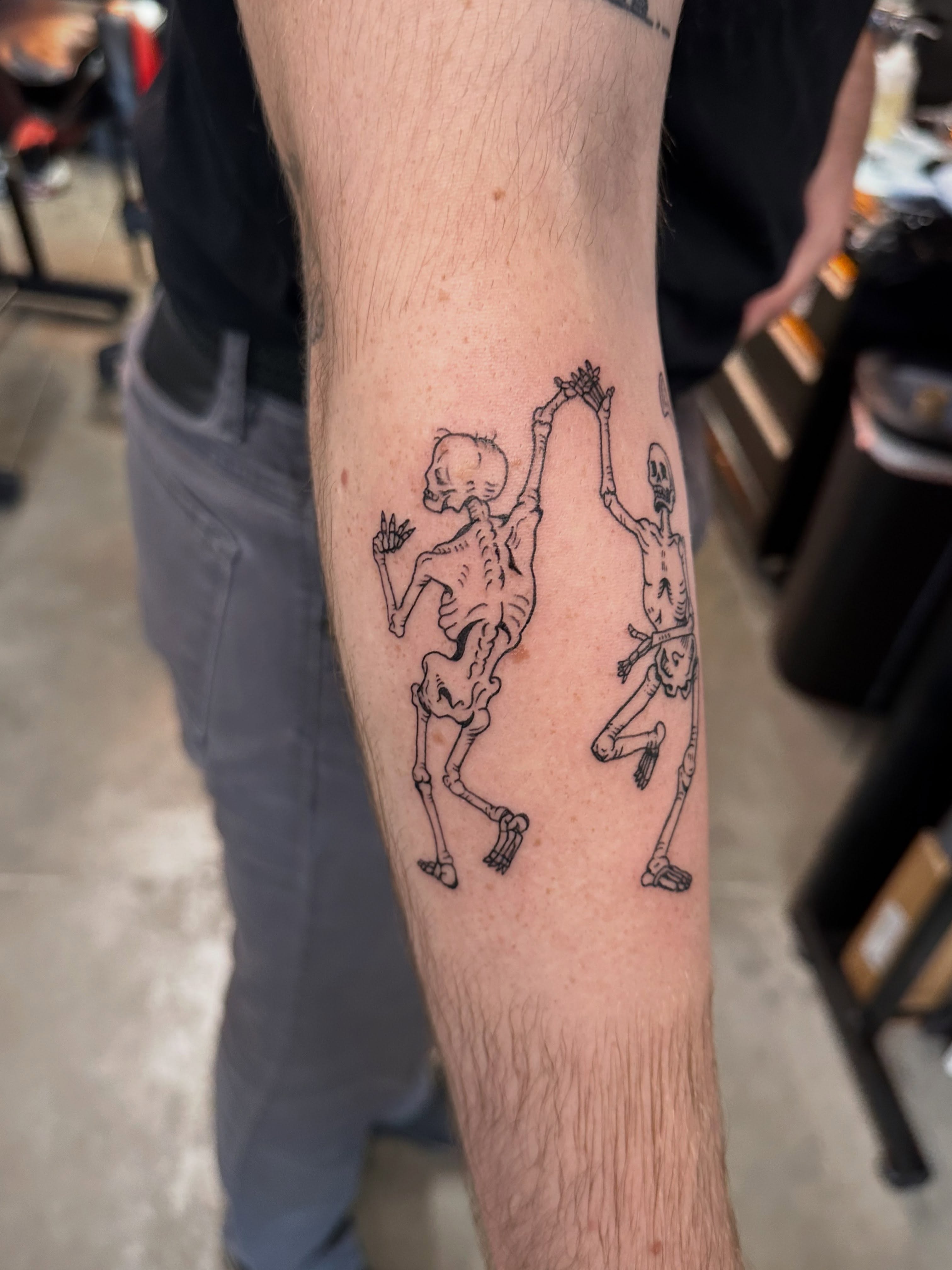 Skeletons dancing black and grey tattoo on the forearm by Galen Thornburg