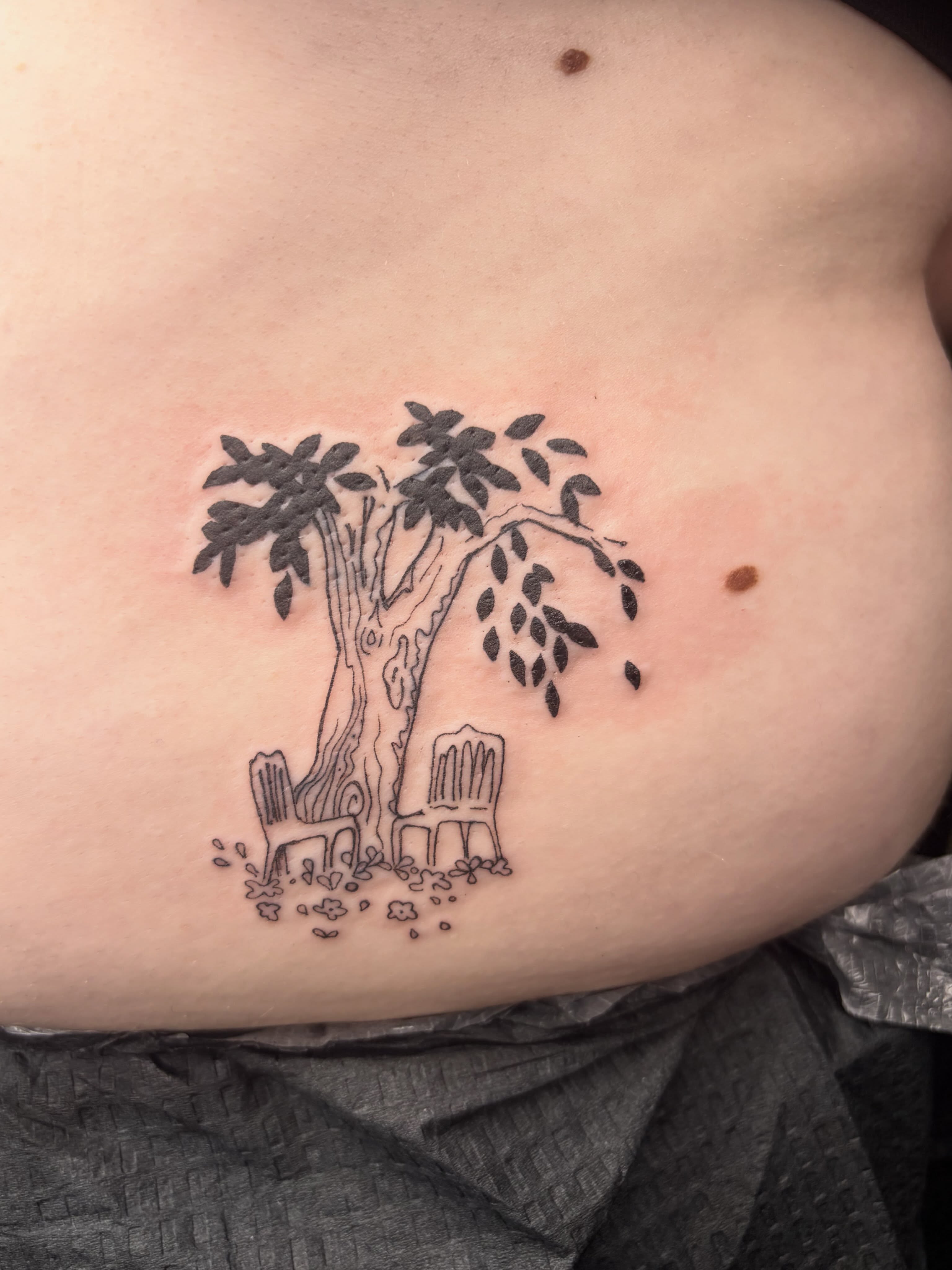 Tree and chairs black and grey tattoo on the ribs by Mattie Robinson