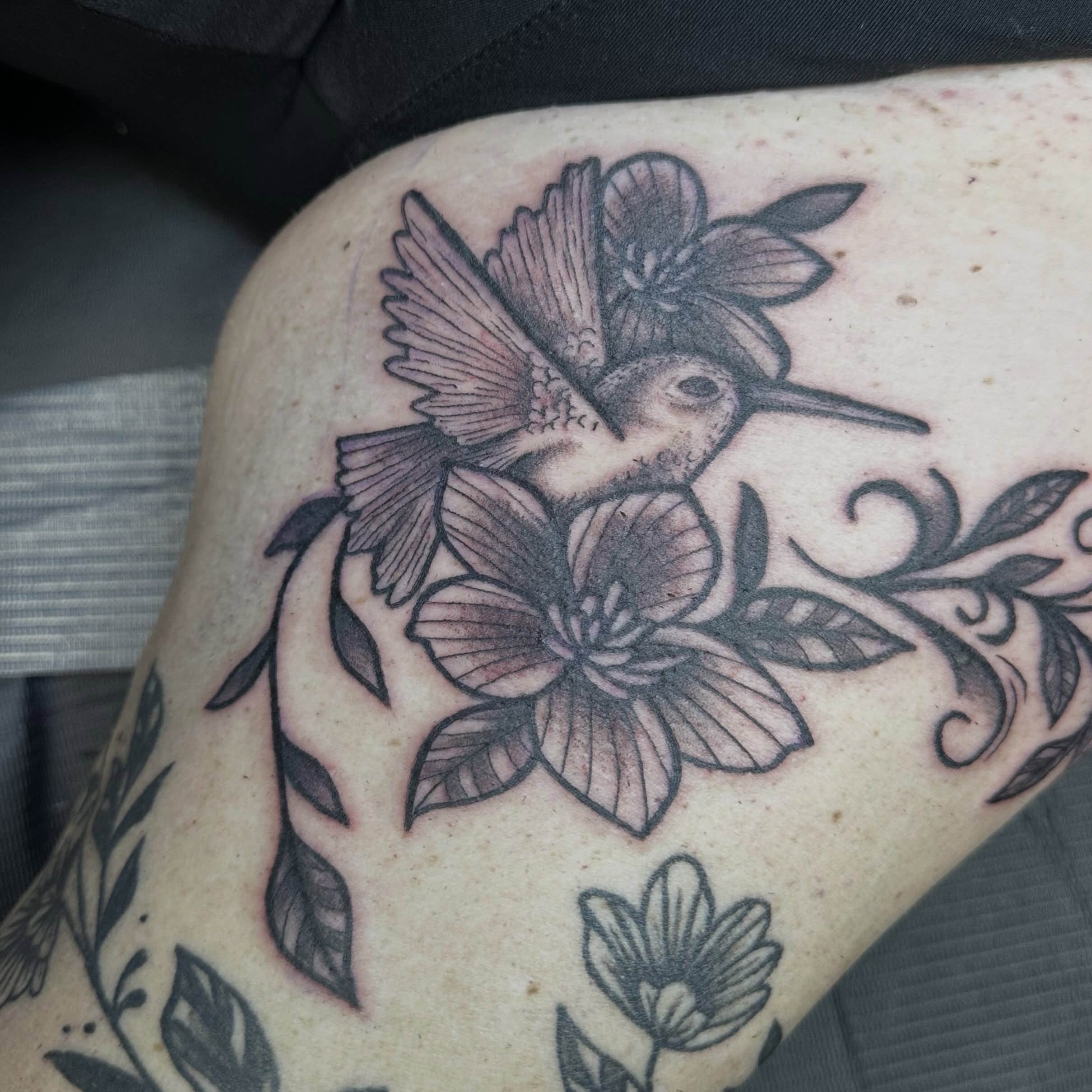Hummingbird and flowers black and grey tattoo on the thigh by Mikaila Jean
