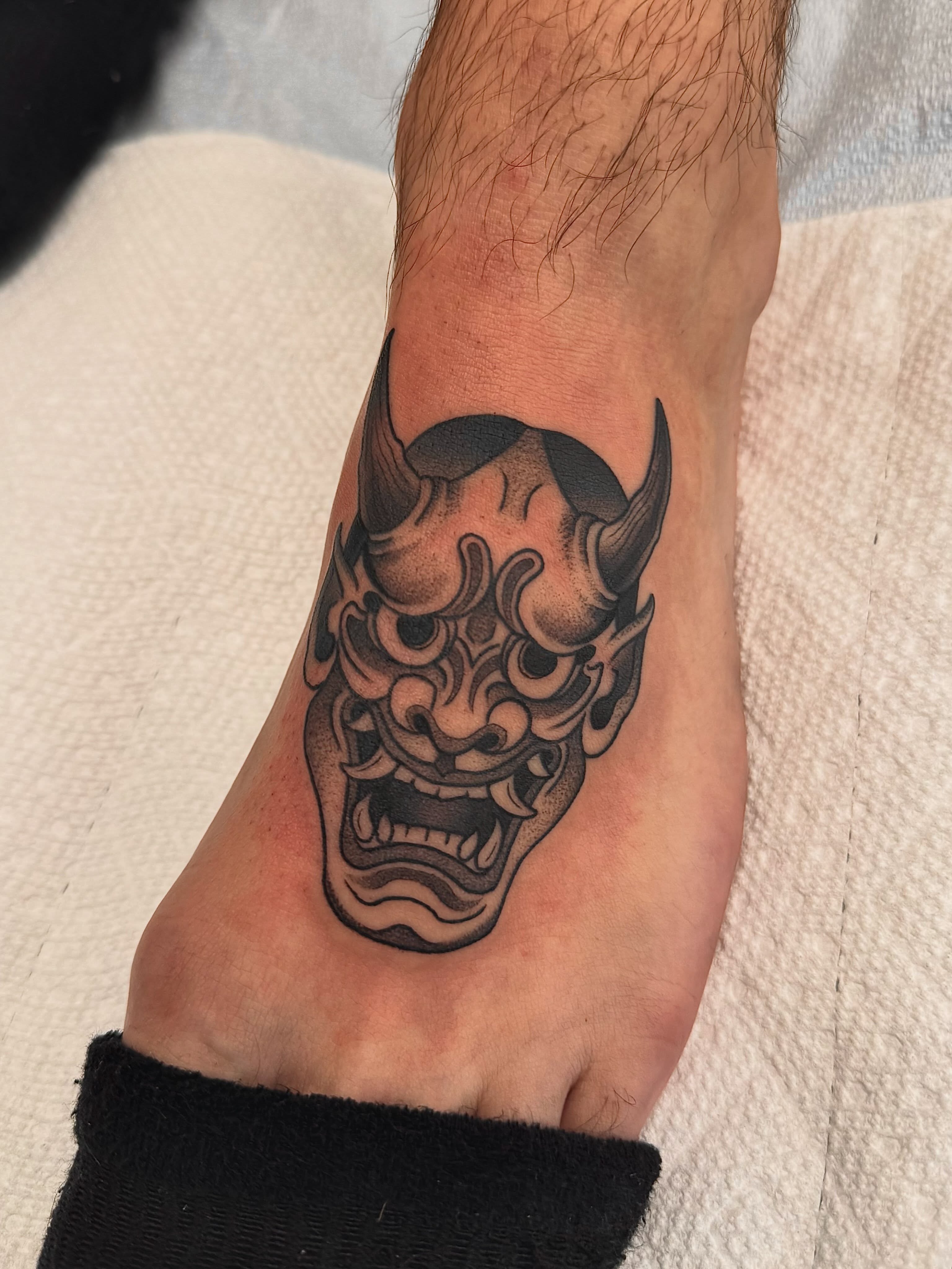 Demon mask black and grey tattoo on the foot by Stefany