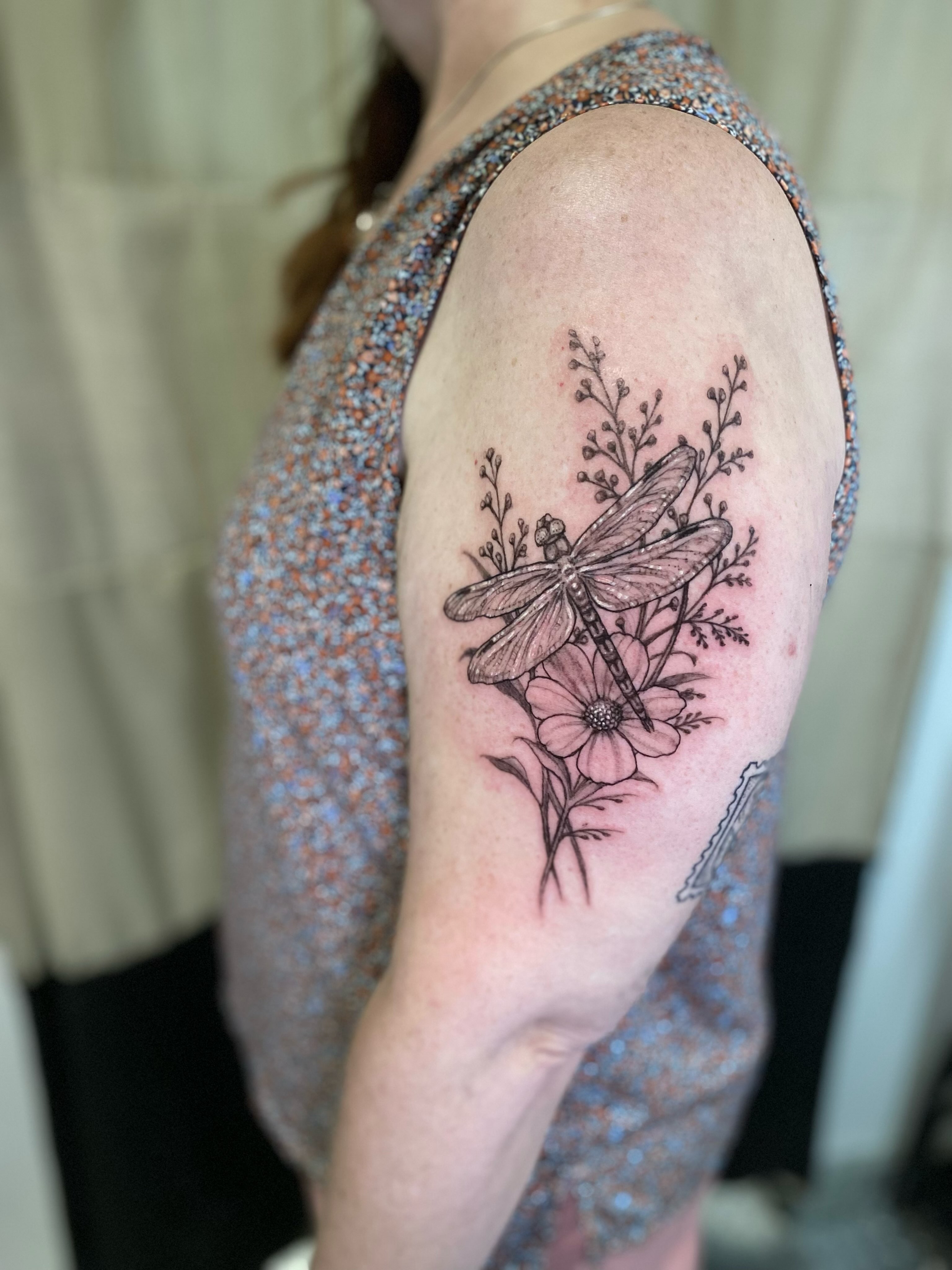 Dragonfly and flowers black and grey tattoo on the upper arm by Dana Jones