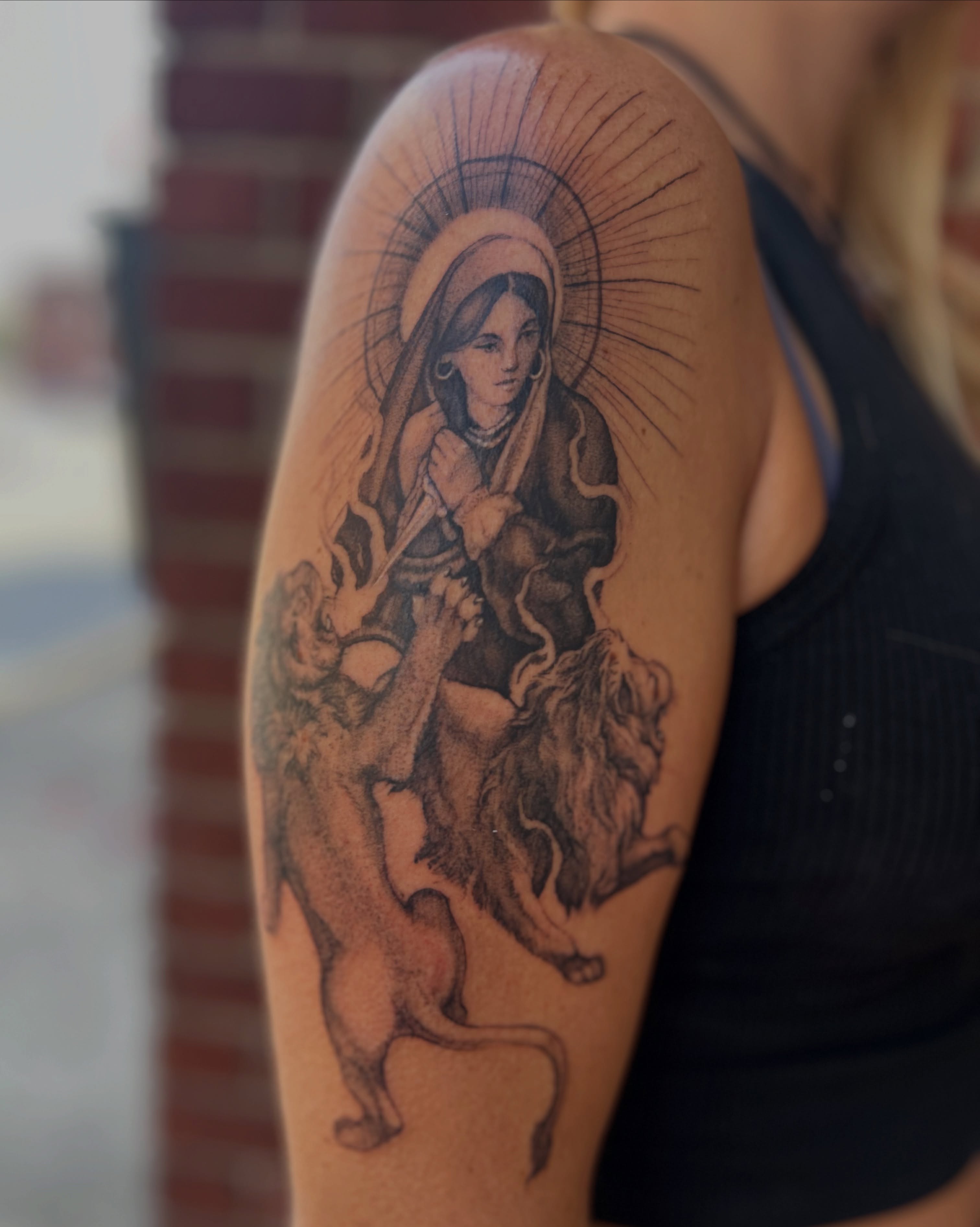 Religious figure with lion black and grey tattoo on the upper arm by Amber Miracle