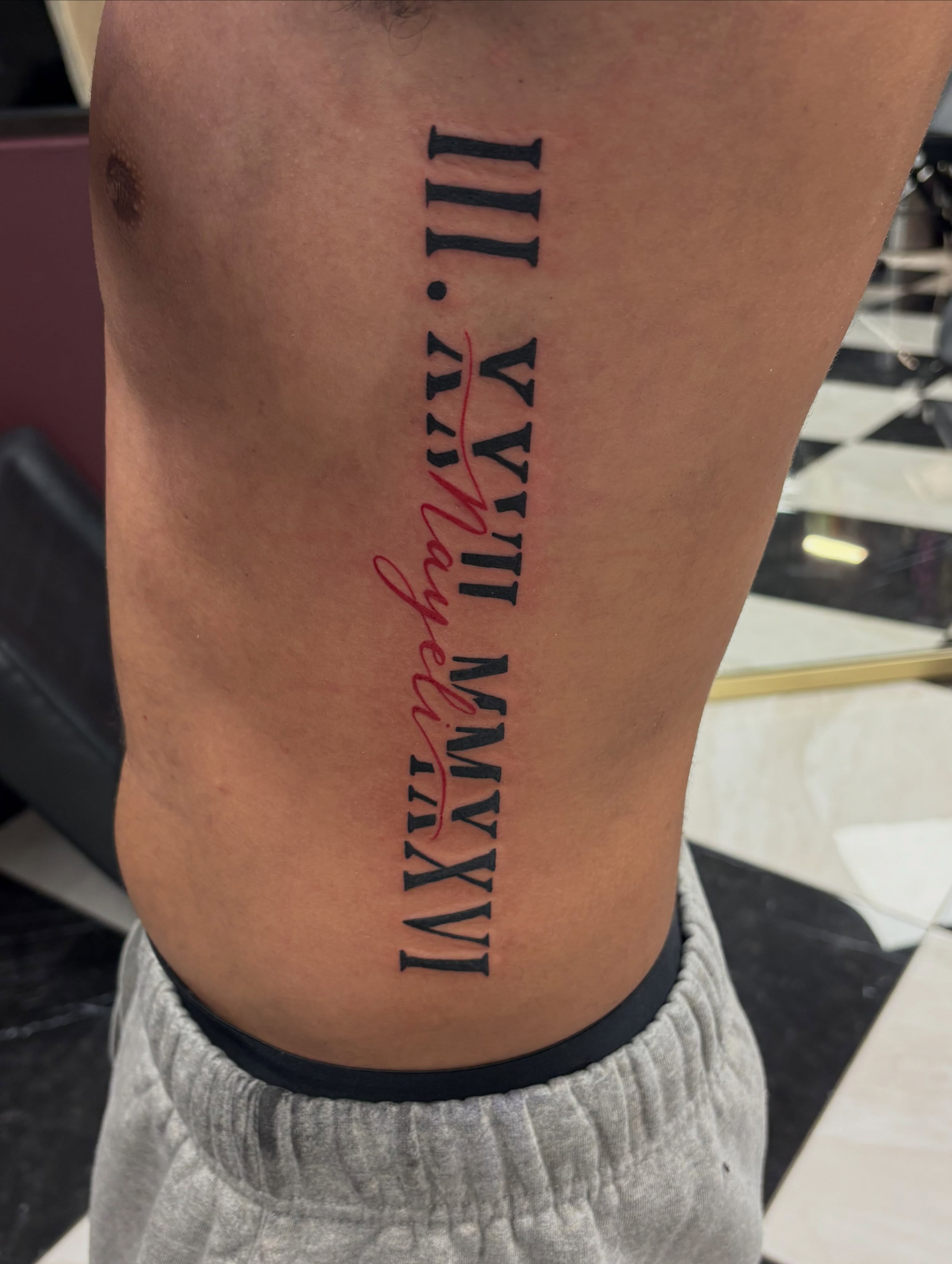 Roman numerals and cursive name lettering script tattoo on the ribs by Kells